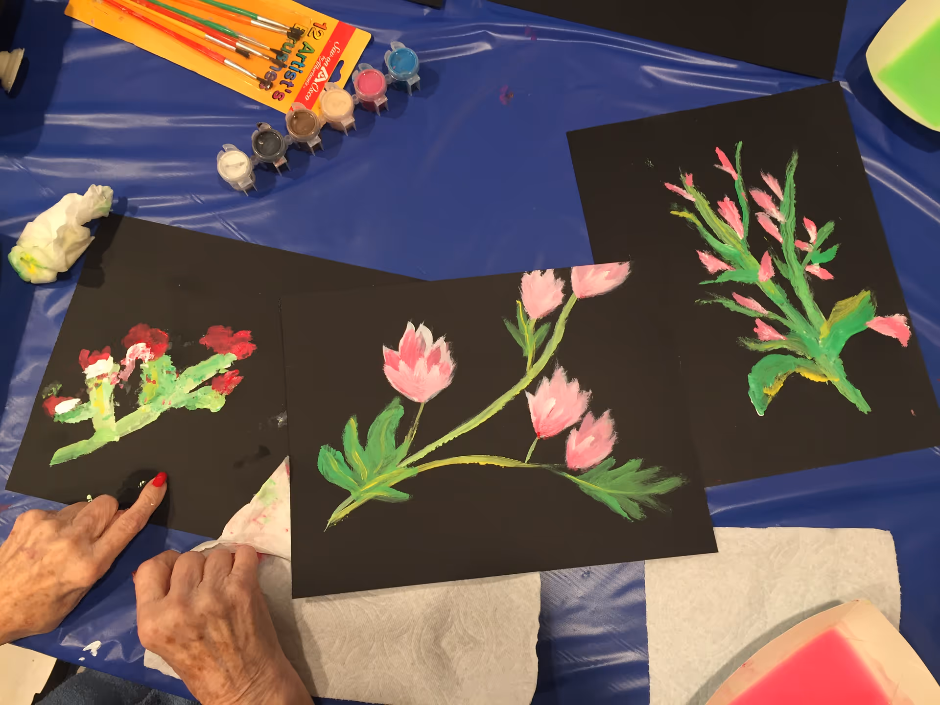 Three black sheets of paper with colorful flower paintings on them placed on a blue tablecloth. A set of small paint containers and paintbrushes are visible at the top left. Two elderly hands with red nail polish are seen at the bottom left, one pointing at a painting and the other resting on a paper towel.