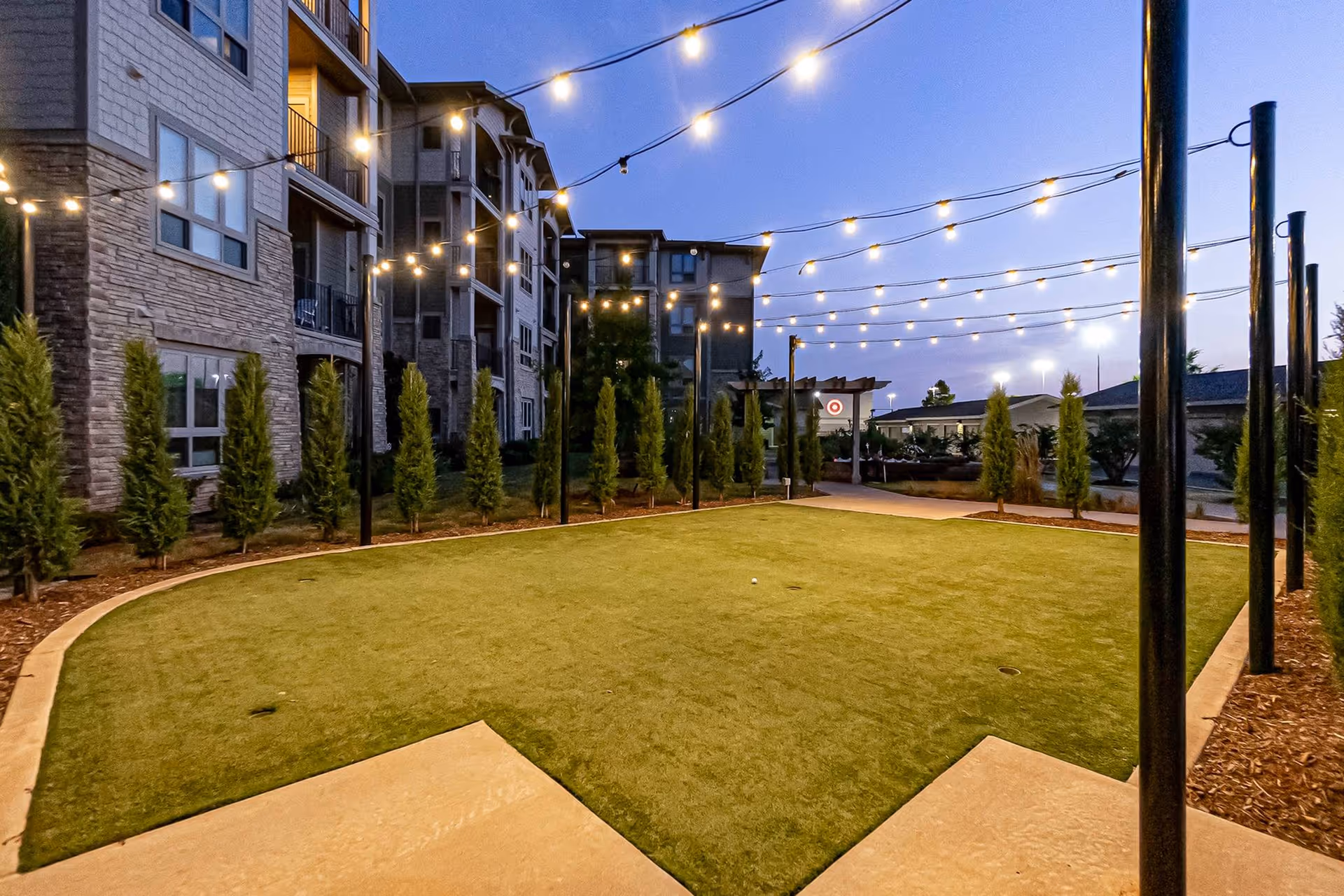 Outdoor area at dusk with string lights hanging above a rectangular putting green surrounded by small trees and a multi-story residential building on the left.