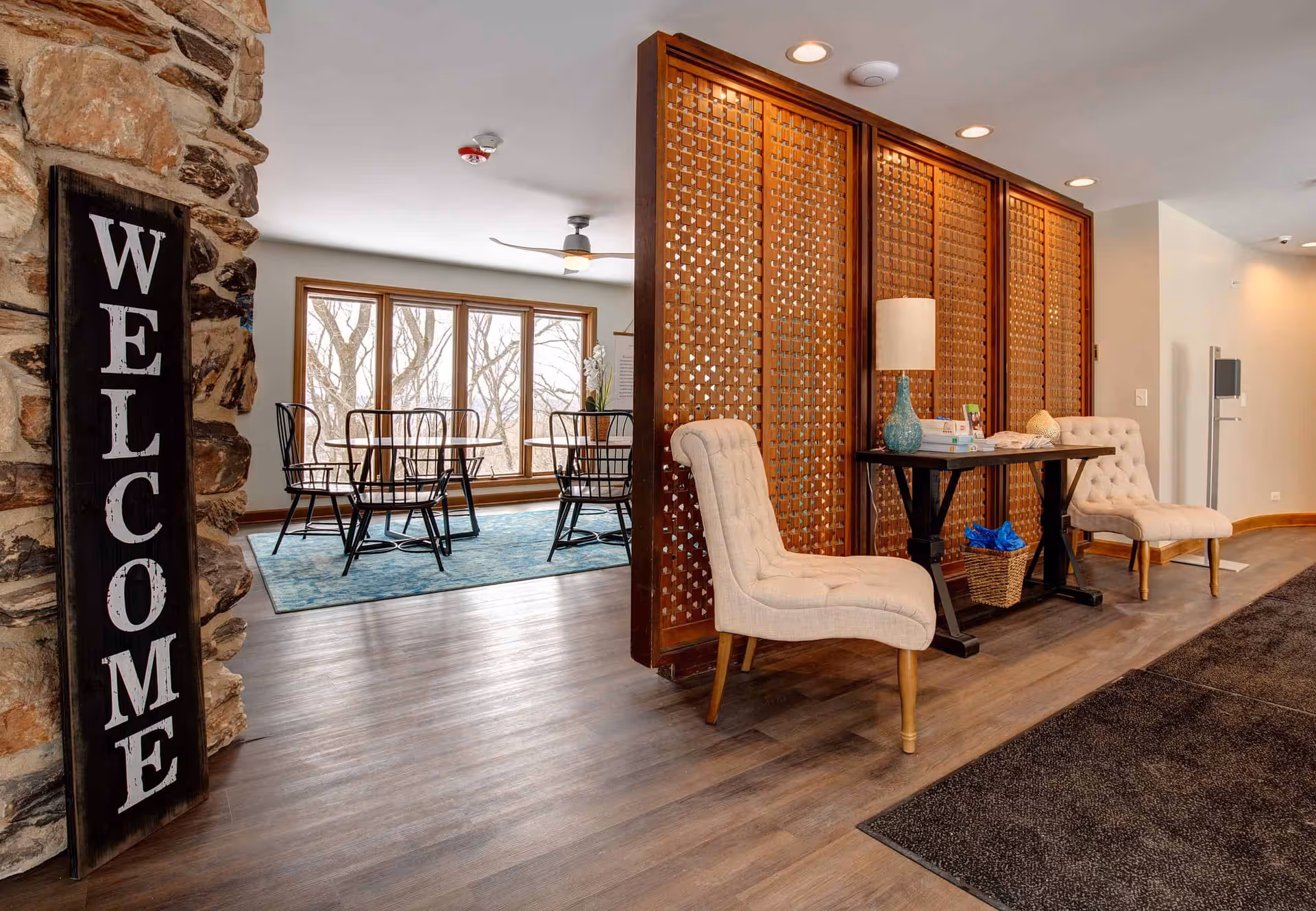 Interior view of a senior living facility showing a welcoming area with a large 'WELCOME' sign on a stone wall. There are two beige tufted chairs and a wooden table with a blue lamp and some items on it. A decorative wooden room divider separates this area from a dining space with a round table and black chairs on a blue rug, next to large windows with a view of leafless trees outside.