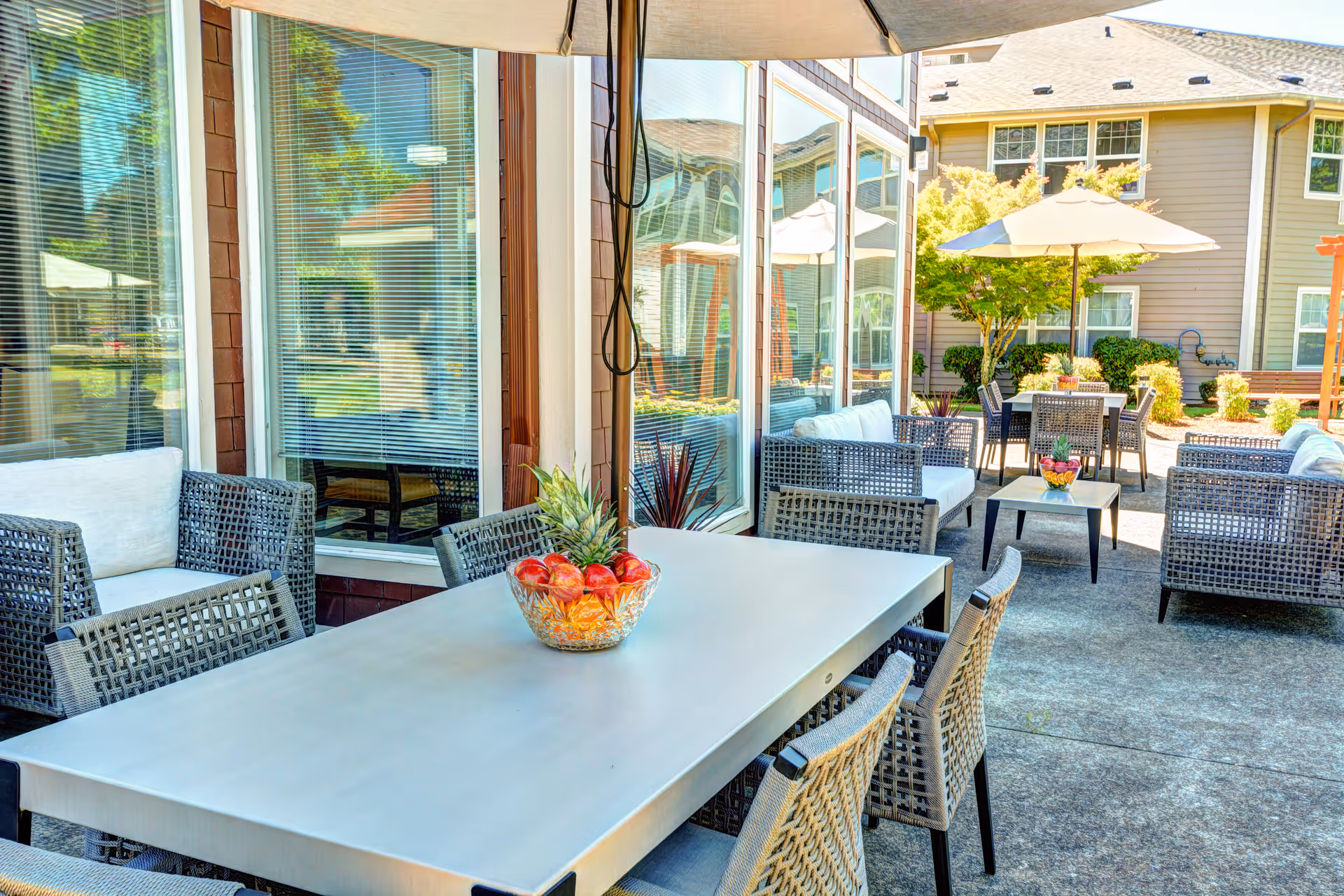 Outdoor patio area at Vineyard Heights Assisted Living featuring a large rectangular table with a bowl of fruit centerpiece, surrounded by woven chairs. Additional seating areas with cushioned chairs and tables are visible under umbrellas. The patio is adjacent to a building with large windows and is surrounded by greenery and trees.