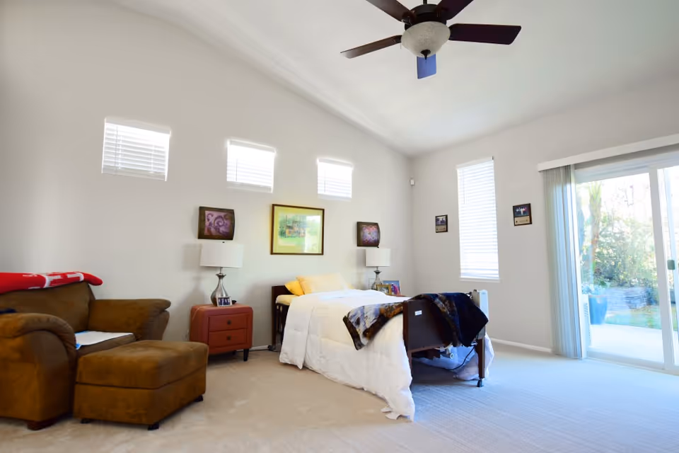 A bright bedroom with a single bed covered in white bedding and a dark blanket at the foot. There is a brown armchair with an ottoman on the left side, a small red nightstand with a lamp, and three small windows high on the wall above the bed. A ceiling fan is mounted on the white ceiling, and a large sliding glass door on the right side opens to an outdoor garden area.