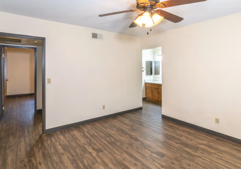 Empty room with wood-patterned flooring, white walls, a ceiling fan with lights, and two doorways leading to other rooms including a bathroom with a wooden vanity and mirror.