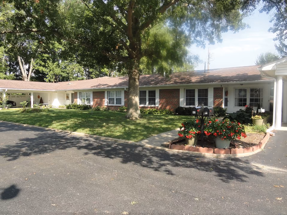 Single-story brick and white siding residential building with multiple windows and a large tree in front. There is a paved driveway and a small garden area with red flowers and two black mailboxes near the entrance.