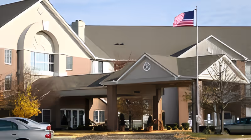Front exterior view of a senior living facility named Danbury North Canton, featuring a covered entrance with columns, an American flag on a flagpole, and a multi-story building with large windows and a peaked roof.