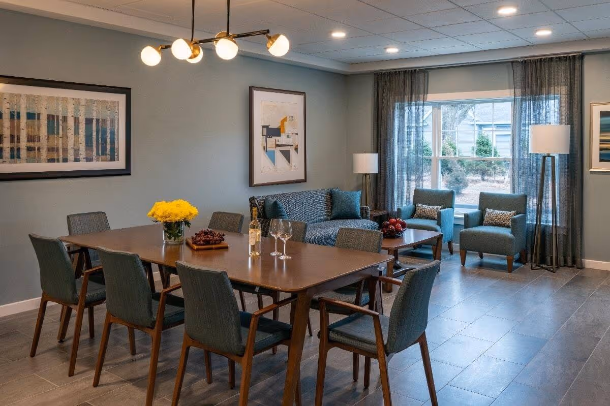 A modern dining and sitting area with a wooden dining table surrounded by eight chairs. On the table, there is a vase with yellow flowers, a bottle of wine, two wine glasses, and a plate of grapes. In the background, there is a blue patterned sofa with two blue cushions, a wooden coffee table with a bowl of red apples, two blue armchairs with patterned cushions, two floor lamps, and large windows with sheer curtains letting in natural light. The walls are painted light blue and decorated with framed artwork.