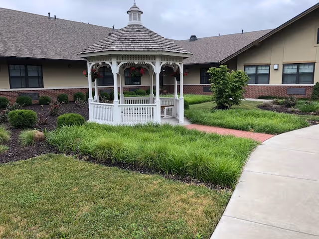 White wooden gazebo in a landscaped courtyard with grass, shrubs, and a one-story brick-and-siding building in the background.