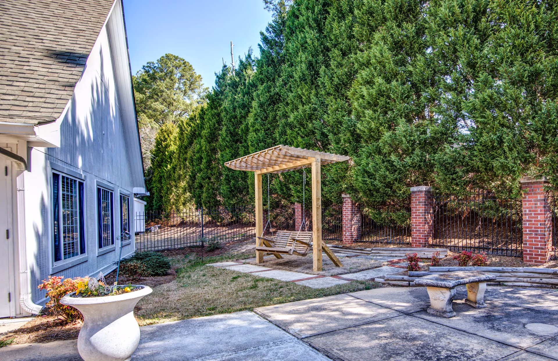 Outdoor area at Oaks at Conyers featuring a wooden swing with a pergola, a concrete bench, a large planter with flowers, and a building with multiple windows. The area is surrounded by tall green trees and a black metal fence with brick pillars.
