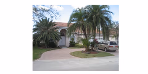 Single-story Mediterranean-style building with arched entry, palm trees and a driveway with a parked minivan.