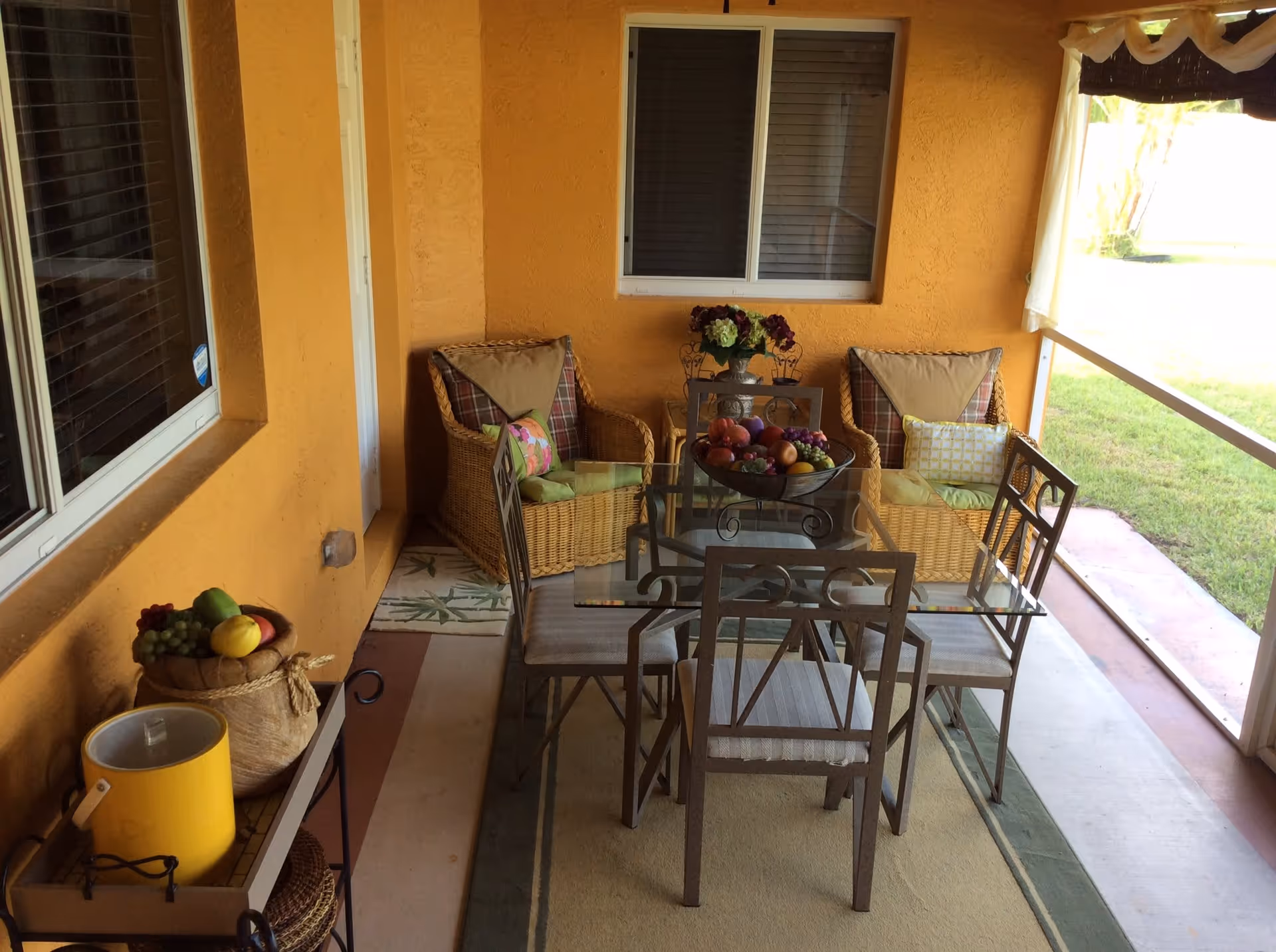 A covered outdoor patio area with a glass-top dining table surrounded by four metal chairs. Behind the table are two wicker armchairs with cushions and pillows. A small side table with a flower vase is positioned between the armchairs. On the left side, there is a small table with a basket of fruit and a yellow container. The patio has orange textured walls, a window with blinds, and a view of green grass outside.