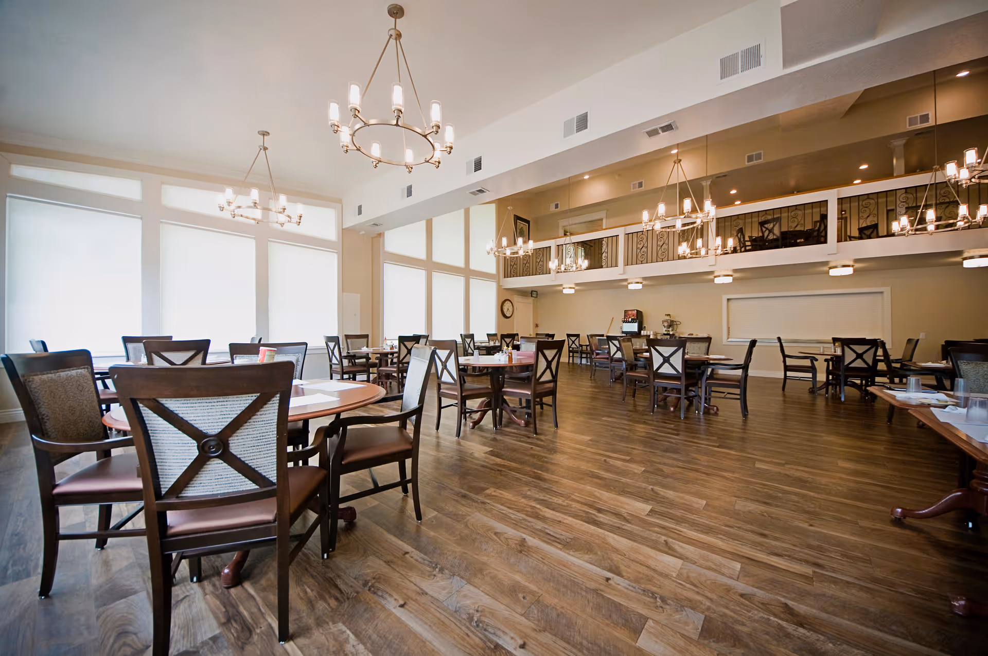 Spacious dining room with multiple round and rectangular wooden tables and chairs arranged neatly on a polished wooden floor. Large windows with white blinds allow natural light to fill the room. Elegant chandeliers hang from the ceiling, and there is a mezzanine level with additional seating visible.