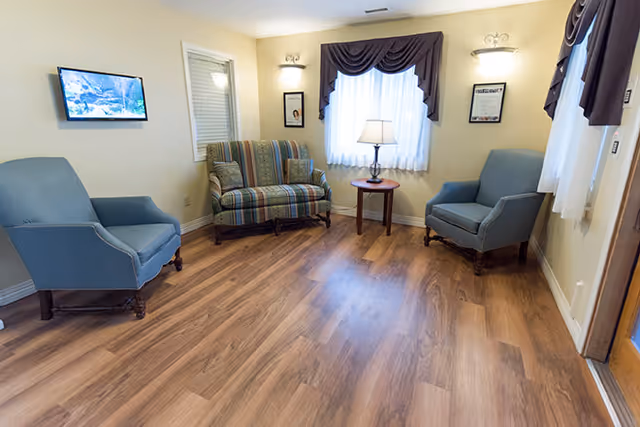 A small sitting room with two blue armchairs, a striped loveseat, a side table with a lamp, wall sconces, and wood-look flooring.