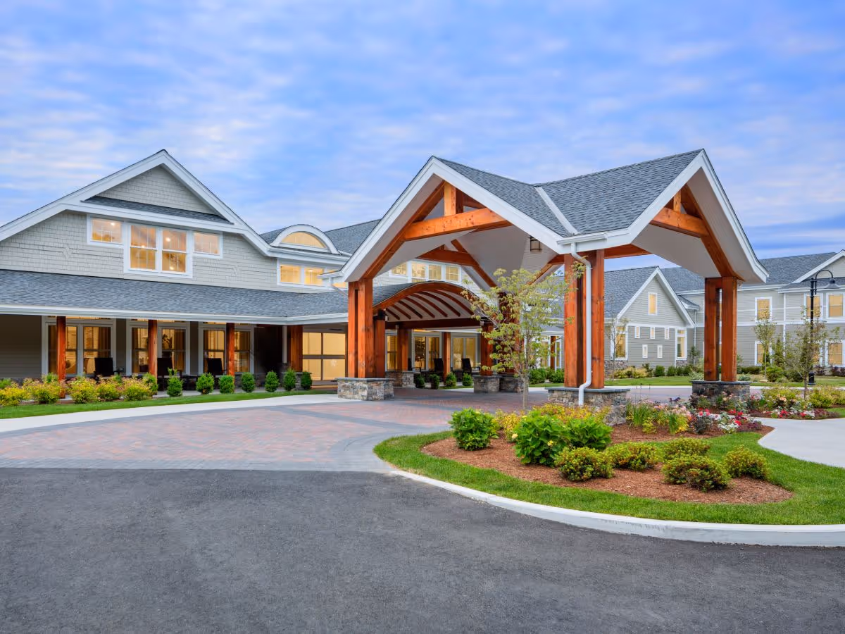 Exterior view of a senior living facility named Maplewood at Brewster, featuring a large covered entrance with wooden beams, landscaped gardens, and a paved driveway under a partly cloudy sky.
