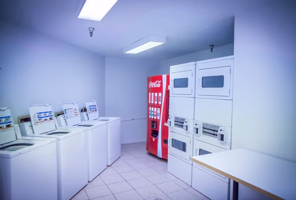 Laundry room with four top-loading washing machines on the left, two stacked dryers on the right, a red Coca-Cola vending machine in the corner, and a white folding table in the foreground.