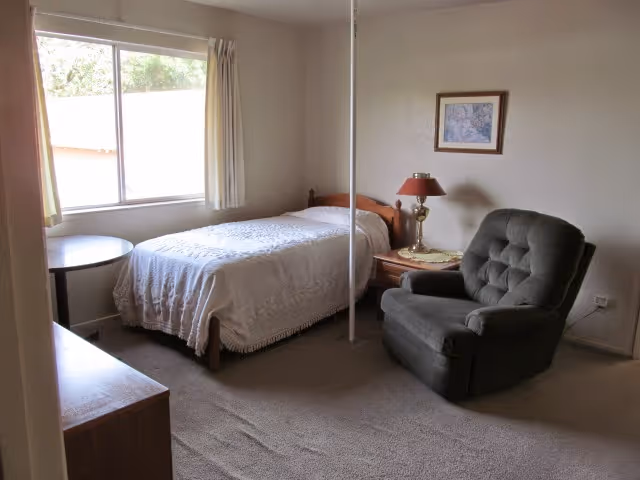 A simple bedroom with a single bed covered with a white bedspread, a wooden nightstand with a lamp, a gray upholstered armchair, a small round table near a window with curtains, and a wooden dresser. The room has beige walls and carpeted floor.