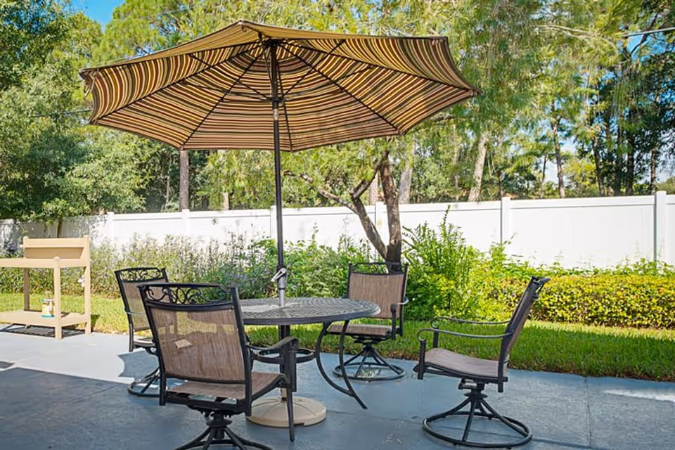 Outdoor patio area with a round metal table and four swivel chairs with mesh backs and seats. A large striped umbrella provides shade over the table. The patio is surrounded by greenery, including bushes and trees, with a white fence in the background.