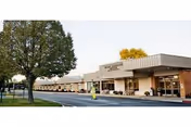 Exterior view of a single-story senior living facility building with a flat roof, large windows, and a covered entrance. There is a tree and a parking lot in front of the building under a clear sky.