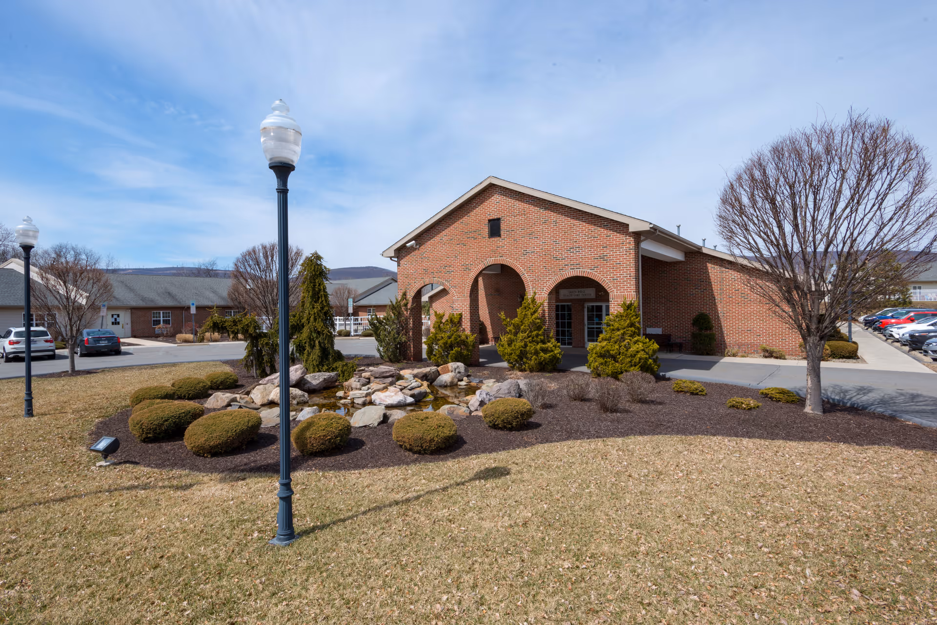 Front entrance of a brick senior living building with an arched entry, landscaped rock garden, lamp posts, and parked cars.