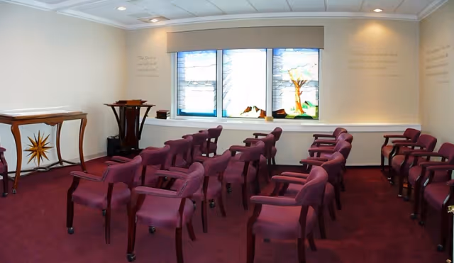 Small chapel-style interior with rows of purple upholstered chairs facing stained-glass windows and a lectern.