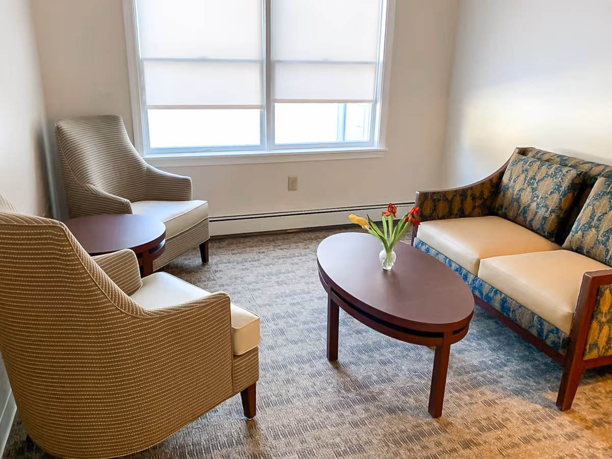 Small sitting area with two upholstered armchairs, a patterned loveseat, an oval coffee table holding a vase of tulips, and a large window.