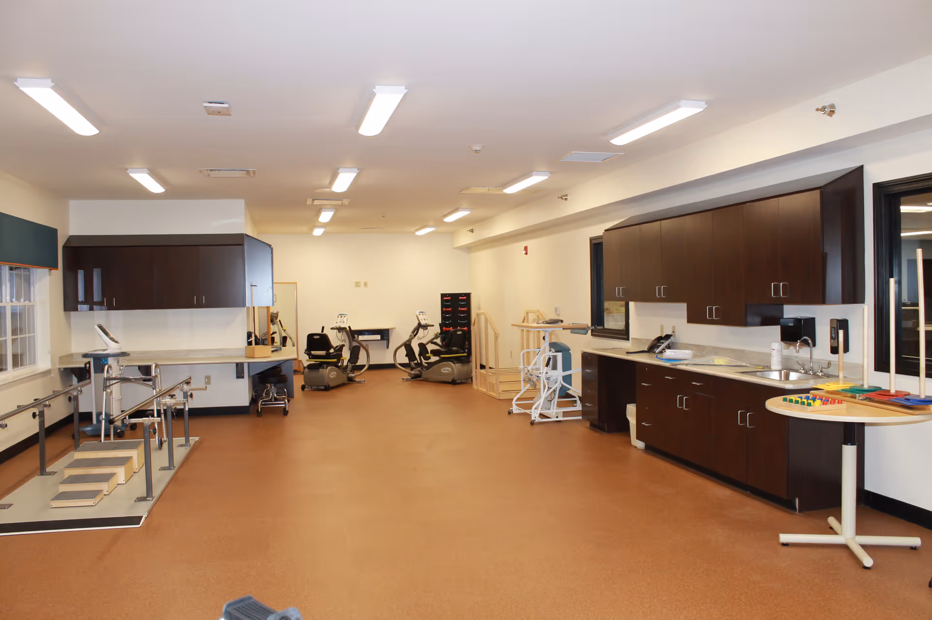 A spacious rehabilitation or physical therapy room with exercise equipment including stationary bikes, parallel bars with stairs for walking practice, and various therapy tools on a round table. The room has brown flooring, white walls, and dark wood cabinets along the right side with a sink and countertop.