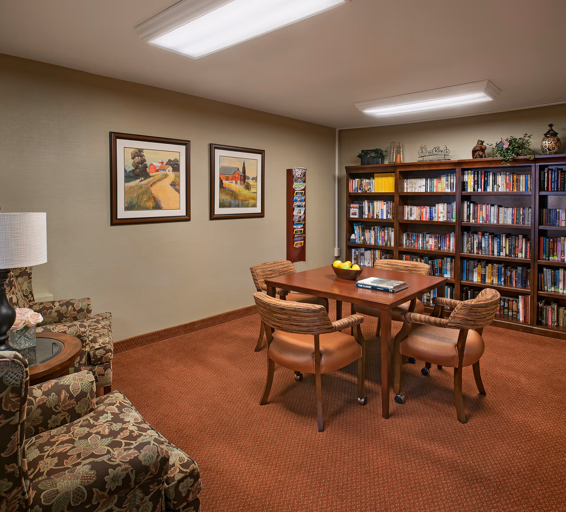 A cozy reading room with a table and four chairs, bookshelves lining the wall, and upholstered armchairs in the corner.