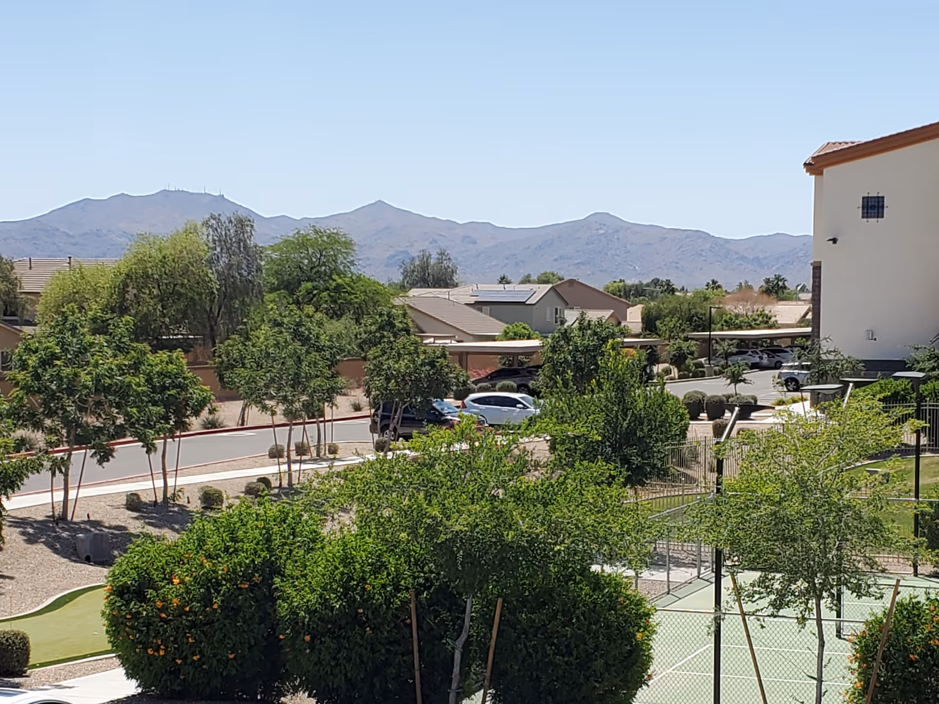 Outdoor view of a senior living facility with green trees, bushes, a small fenced area, parked cars, residential buildings, and mountains in the background under a clear blue sky.