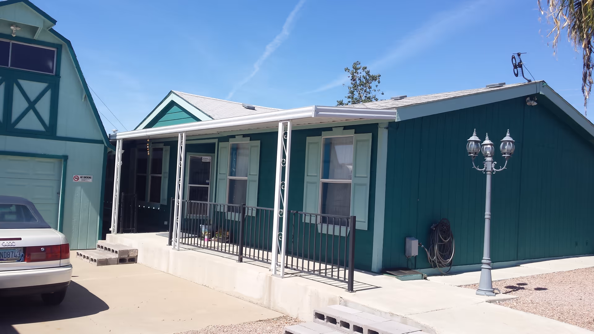 Exterior view of a teal-colored assisted living facility building with a covered porch, black railing, and two windows with light green shutters. A white car is parked on the left side near a garage with a 'No Smoking' sign. There is a vintage-style street lamp on the right side and a clear blue sky above.