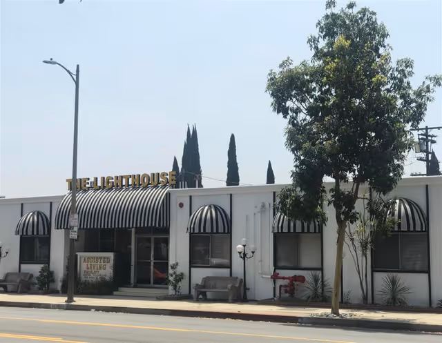Exterior view of The Lighthouse assisted living facility with a white building featuring black and white striped awnings over windows and entrance. There are benches and a tree in front of the building, and a street lamp is visible on the sidewalk.