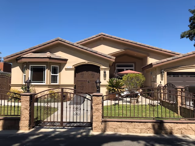 Front exterior view of a single-story beige stucco building with a tiled roof, decorative wrought iron gate and fence, a concrete pathway leading to a large wooden double door, and a small garden area with plants and a red patio umbrella.