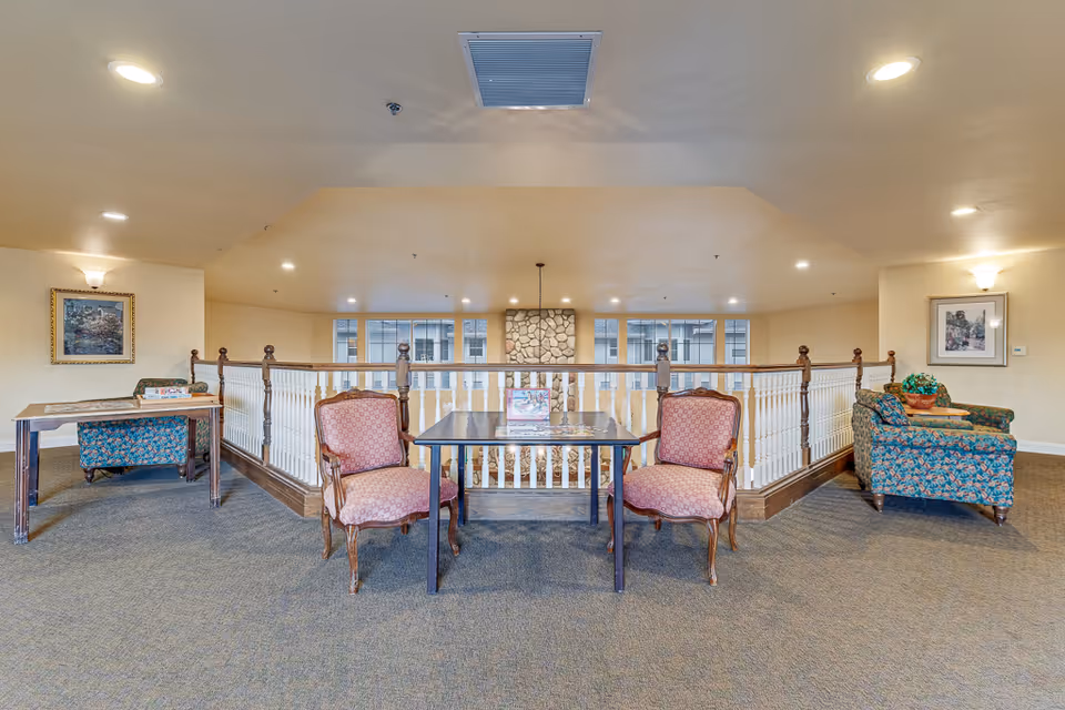 Upstairs common sitting area with upholstered chairs and tables arranged along a wooden railing overlooking a stone fireplace.