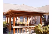 A wooden gazebo with a shingled roof situated in an outdoor courtyard area surrounded by shrubs and plants, with a building visible in the background.