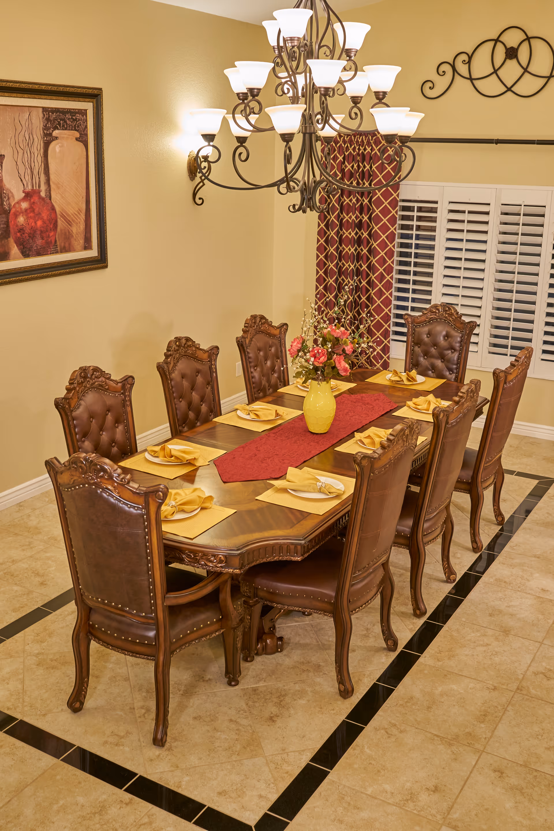 A formal dining room with a long wooden table surrounded by eight ornate brown leather chairs. The table is set with yellow napkins on placemats and a red table runner with a yellow vase holding a floral arrangement in the center. A chandelier with multiple lights hangs above the table. The room has beige walls, a framed painting, a window with white shutters, and red patterned curtains.