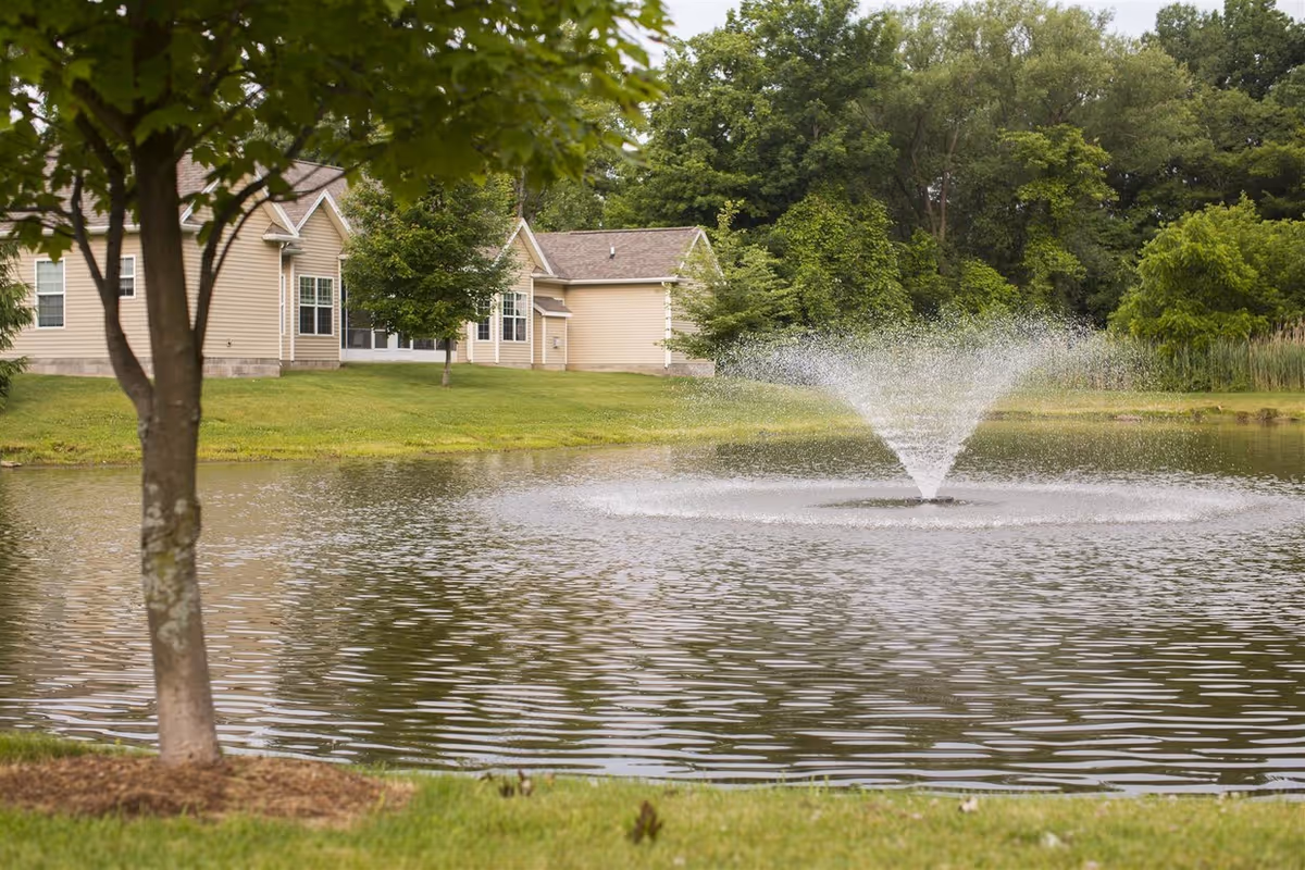 A pond with a central fountain in front of beige townhomes surrounded by trees and a grassy lawn.
