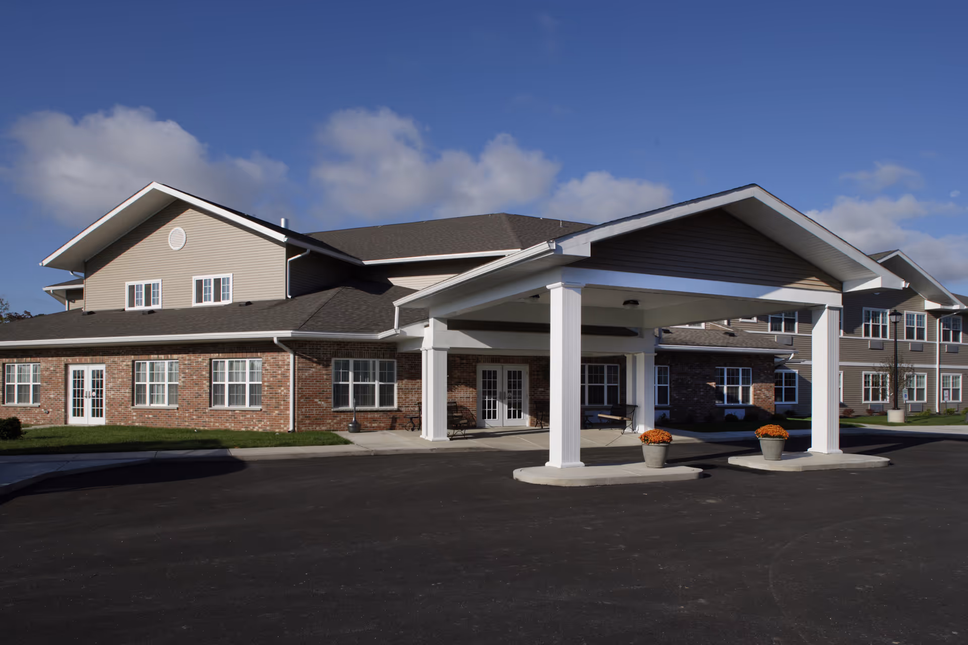 Exterior view of a senior living facility building with a covered entrance supported by white columns, brick and siding facade, multiple windows, and a clear blue sky with some clouds.