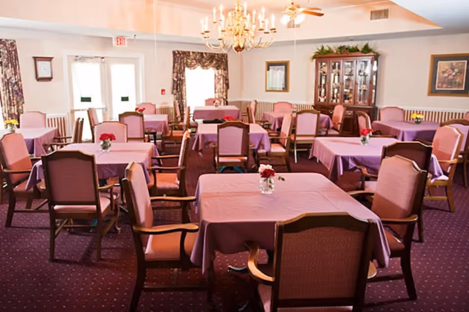 A dining room with multiple square tables covered with purple tablecloths, each table surrounded by wooden chairs with pink cushions. Small flower vases with red and yellow flowers are placed on the tables. The room has a chandelier hanging from the ceiling, floral curtains on the windows, a clock on the wall, and a wooden cabinet with glass doors displaying decorative items.