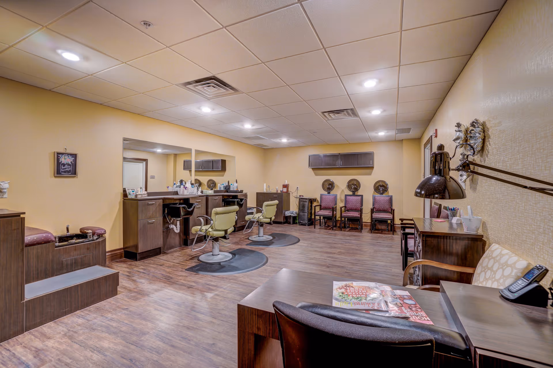 Interior view of a salon area in a senior living facility with two green salon chairs in front of mirrors, a pedicure station with a foot bath, several chairs lined up against the wall, and a desk with a lamp and magazines.
