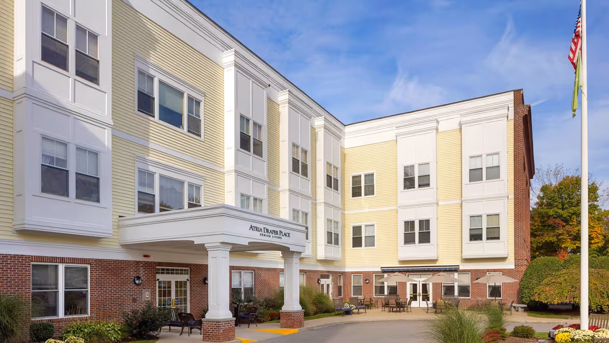Exterior view of Atria Draper Place senior living facility showing a three-story building with a combination of yellow siding and red brick. There is a covered entrance with white columns and outdoor seating areas with tables and chairs. A flagpole with an American flag is visible on the right side, and there are trees and bushes around the building under a partly cloudy blue sky.