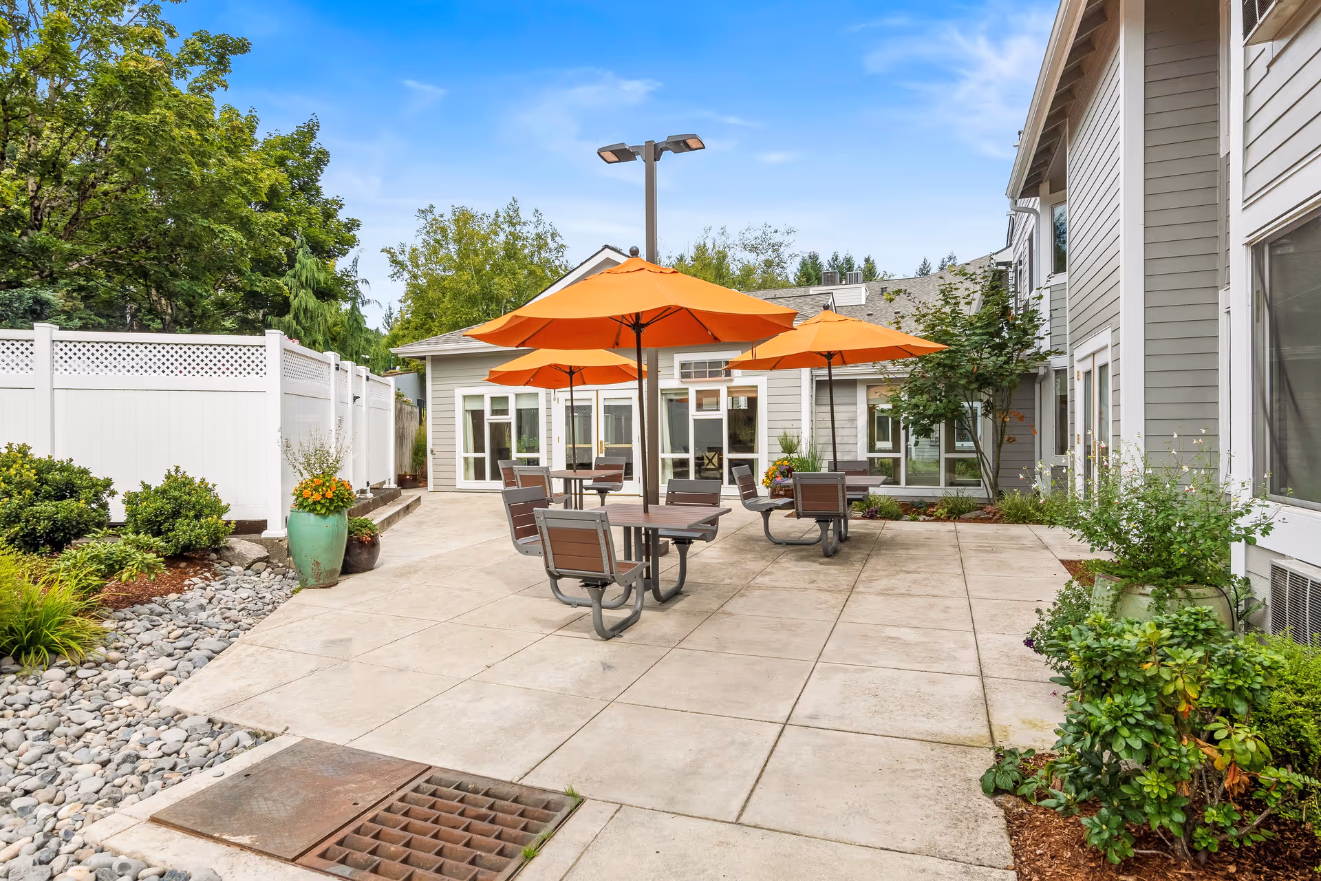 Outdoor patio area at Regency Park featuring several tables with orange umbrellas, surrounded by greenery and a white fence, adjacent to a light gray building under a blue sky.