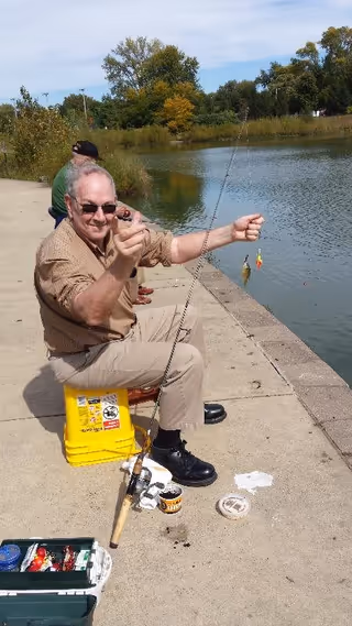 An elderly man wearing sunglasses and a brown shirt is sitting on a yellow bucket by the edge of a pond, holding a fishing rod with a small fish caught on the line. Another person is sitting behind him, also fishing. The scene is outdoors with trees and grass in the background under a partly cloudy sky.