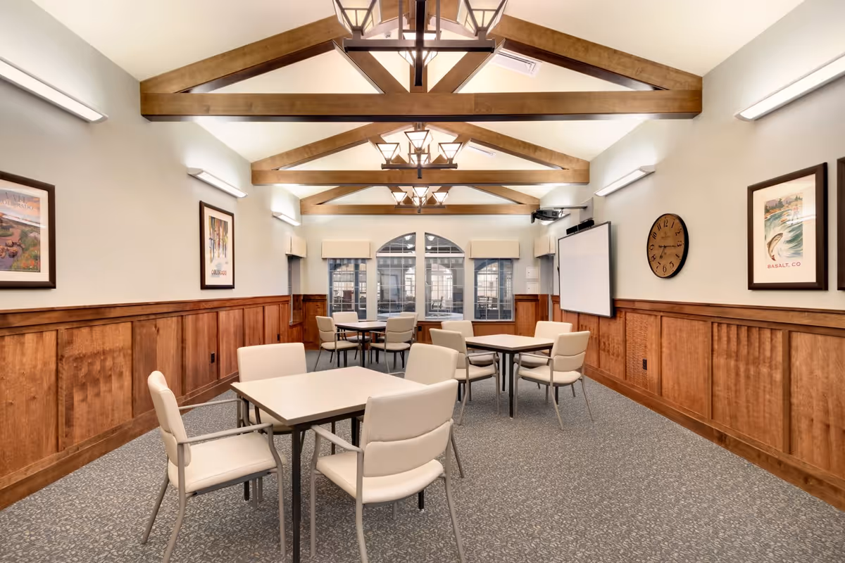 A well-lit room with wooden beams on the ceiling and multiple square tables surrounded by beige cushioned chairs. The walls have wood paneling on the lower half and light-colored paint above, decorated with framed artwork and a round clock. There is a projector and a large screen on one wall, and windows with beige valances at the far end of the room.