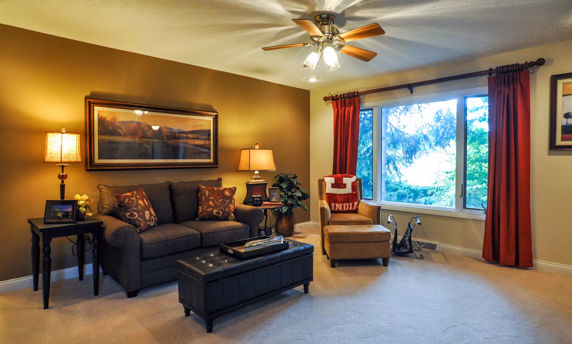 A cozy living room with a dark gray sofa adorned with patterned pillows, a black tufted ottoman in front, and a beige armchair with a red and white blanket draped over it. The room has warm lighting from two table lamps on either side of the sofa, a ceiling fan with lights, a large window with red curtains showing greenery outside, and framed artwork on the walls.