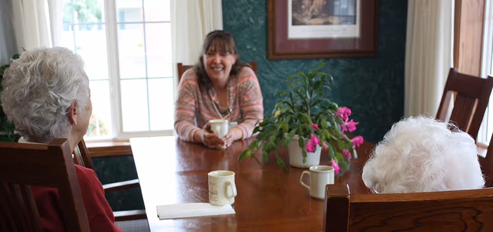 Three people—two older adults seen from behind and a smiling woman—sit around a wooden table with mugs and a potted plant in a bright dining area.