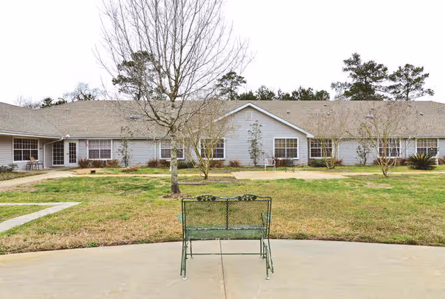A green metal bench on a concrete patio facing a lawn and a one-story light-gray residential building with leafless trees.