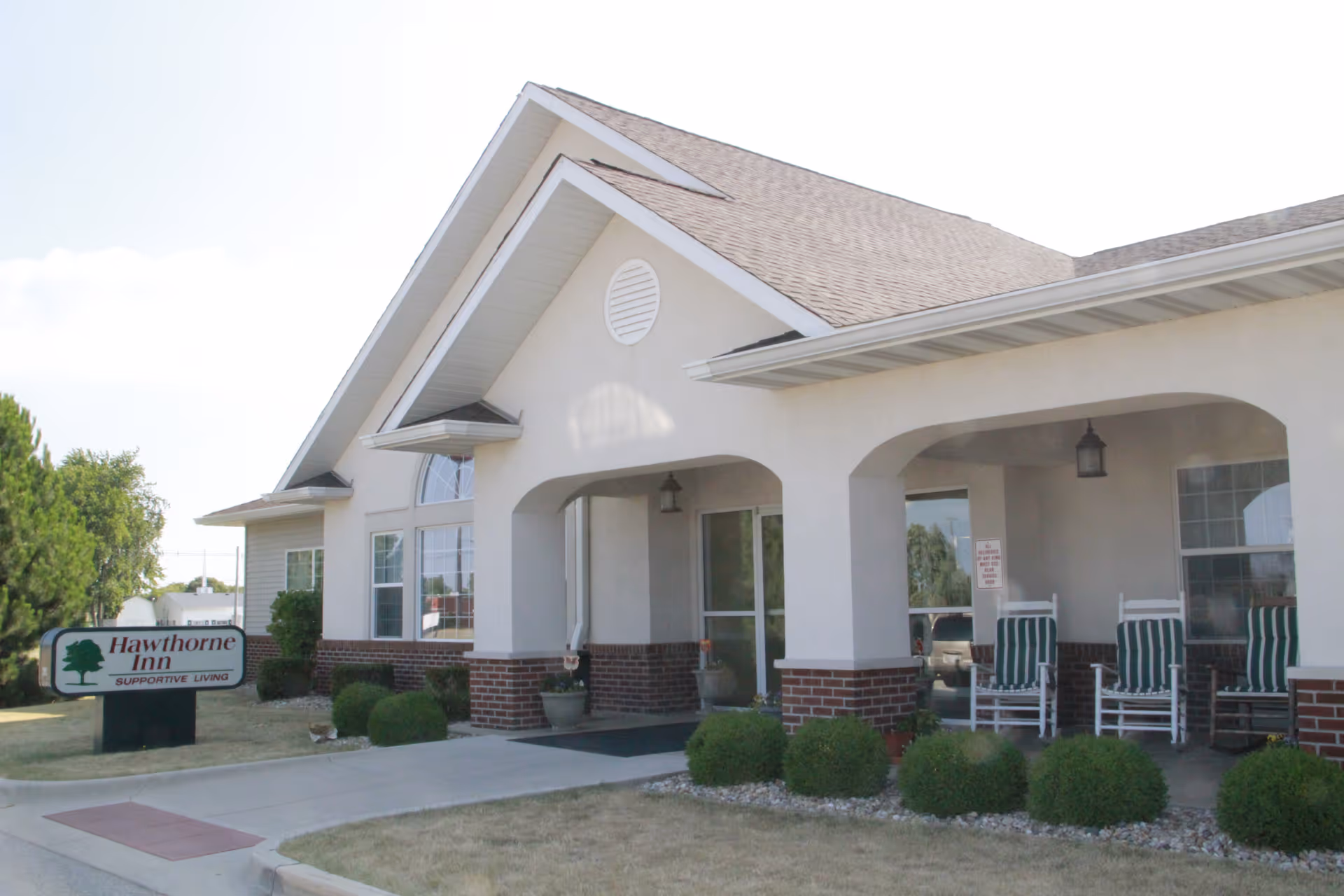 Exterior view of a single-story supportive living facility named Hawthorne Inn, featuring a covered entrance with white pillars, brick accents, several green and white striped rocking chairs on the porch, and neatly trimmed bushes along the walkway.