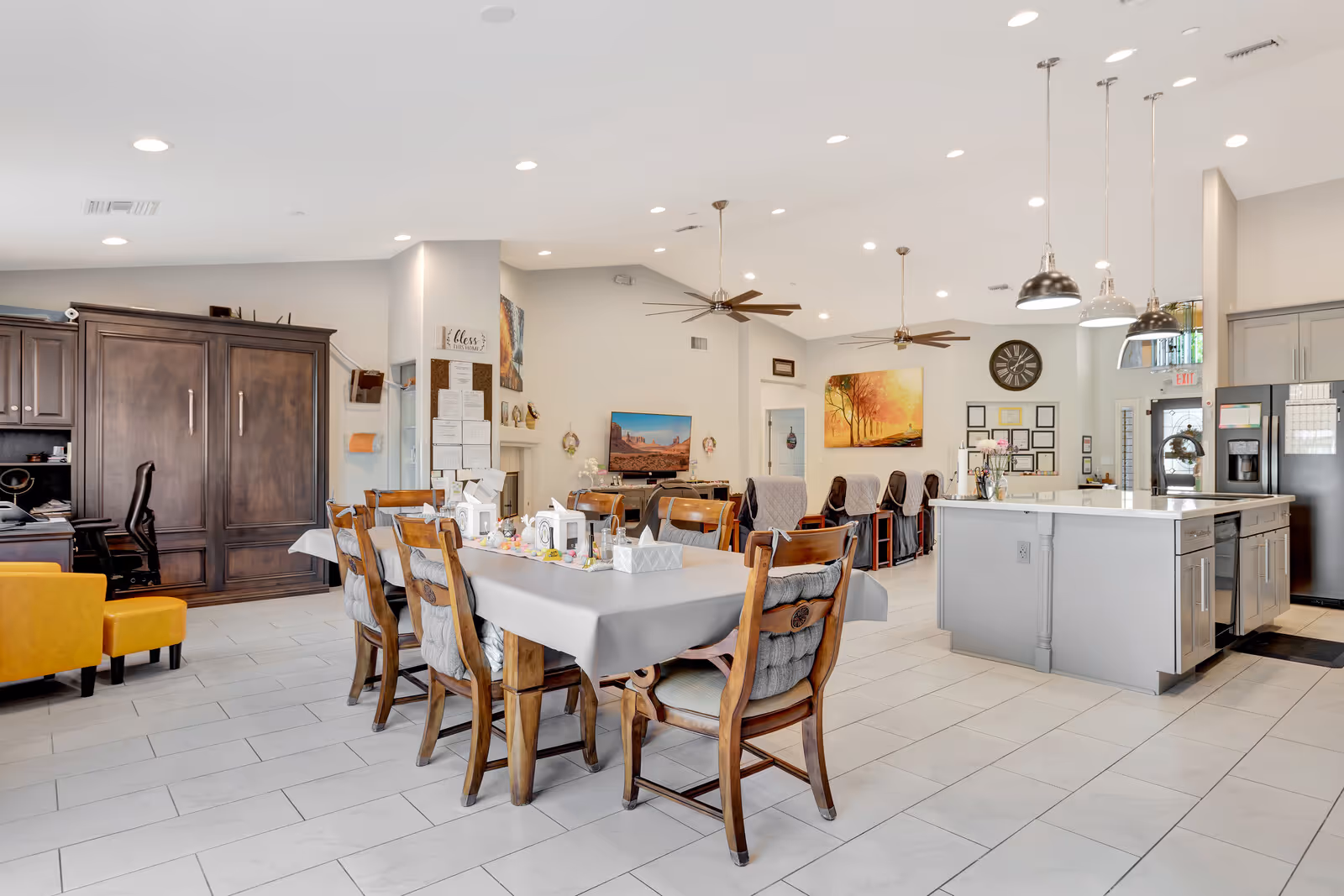 A spacious, well-lit common area in a senior living facility featuring a large dining table with wooden chairs, a modern kitchen island with pendant lights, comfortable recliner chairs lined up facing a wall-mounted TV, and decorative wall art. The room has white tiled flooring, ceiling fans, and a cozy, welcoming atmosphere.