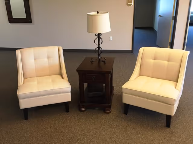 Two beige upholstered armchairs with dark wooden legs positioned on either side of a dark wooden side table with a drawer and a decorative lamp on top, set against a beige wall and carpeted floor in an interior room.