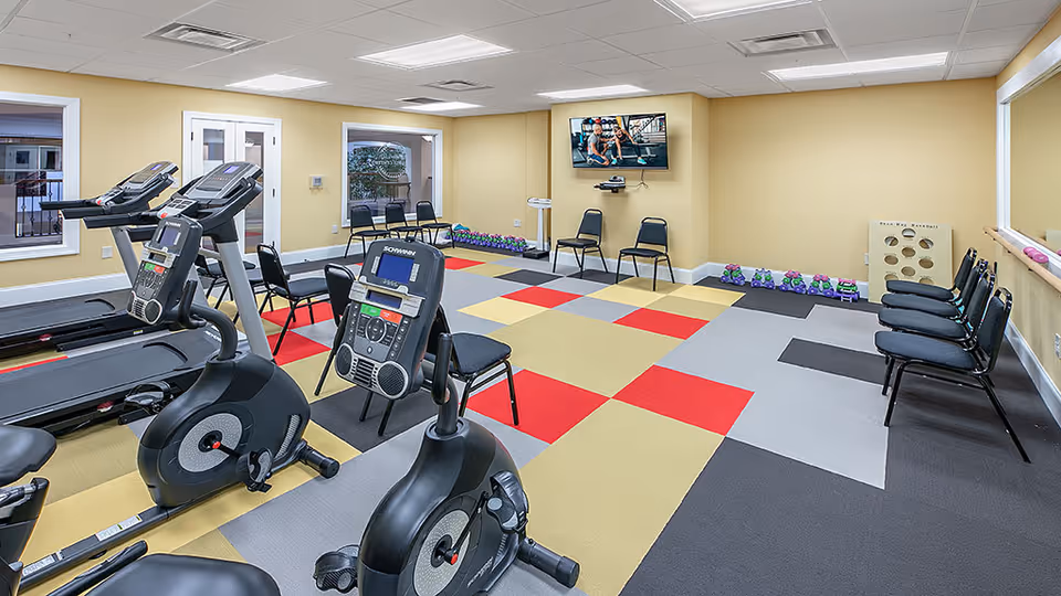 A senior living facility exercise room with stationary bikes, treadmills, chairs arranged along the walls, a television mounted on the wall showing an exercise video, colorful checkered flooring, and various small exercise equipment like dumbbells and a balance board.
