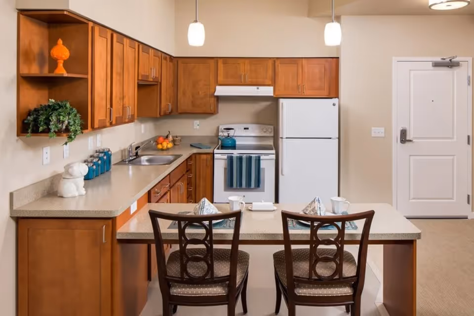 A kitchen area in a senior living facility with wooden cabinets, a white refrigerator, stove, and sink. There is a countertop with two chairs set with napkins and cups. The walls are beige, and two pendant lights hang from the ceiling. A white door is visible in the background.
