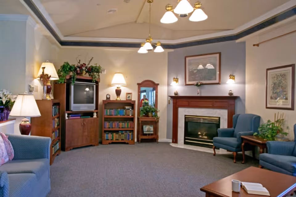 A cozy living room with blue upholstered chairs and a sofa, a wooden coffee table with a book and a cup, a fireplace with a wooden mantel, framed artwork on the walls, several lamps providing warm lighting, bookshelves filled with books, a TV on a wooden stand, and various plants adding greenery to the space.