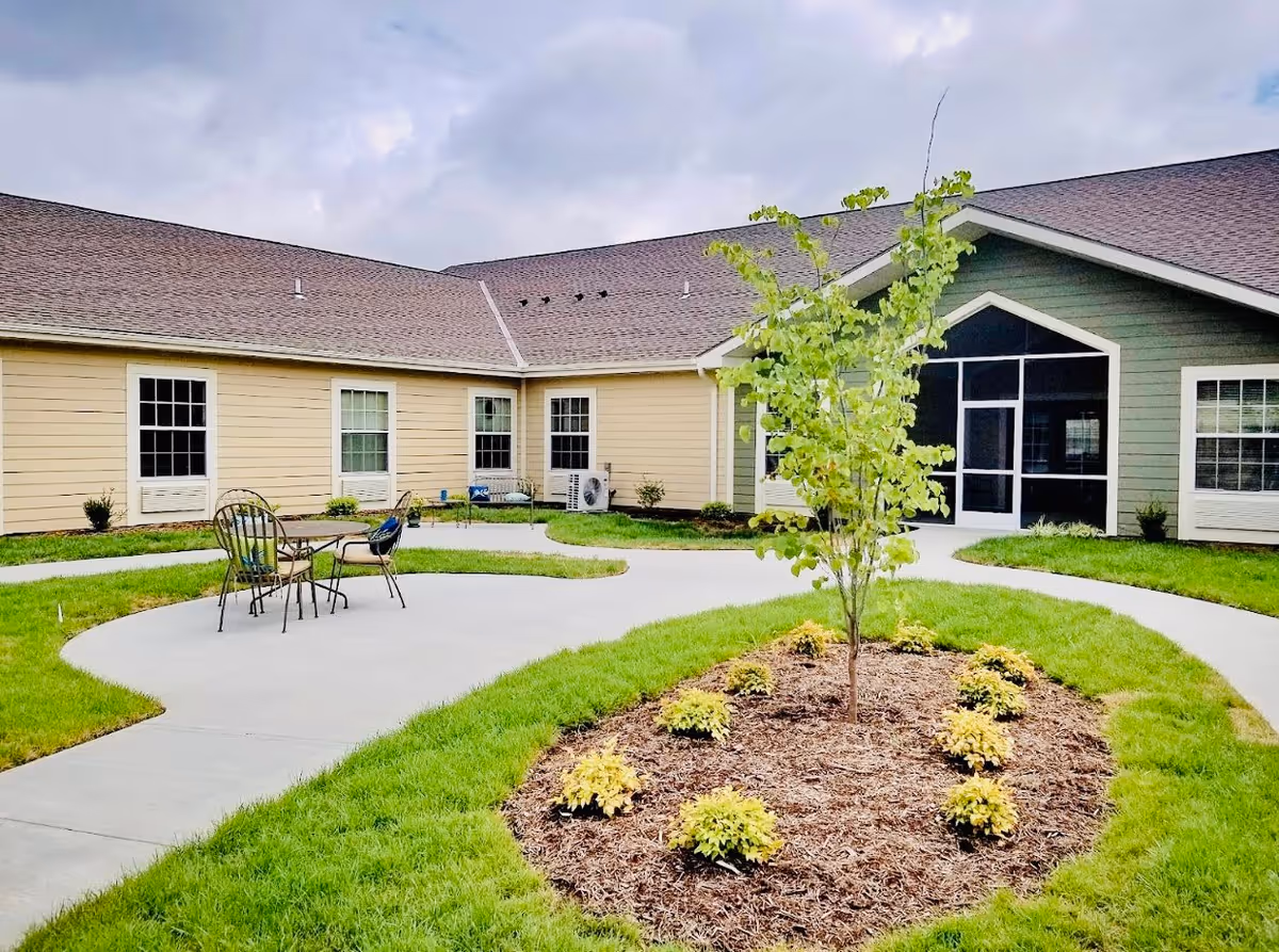 Outdoor courtyard area of a senior living facility with a small tree and plants in a mulched bed surrounded by green grass. There is a concrete walkway with a round table and chairs. The building has beige and green siding with multiple windows and a screened porch entrance.